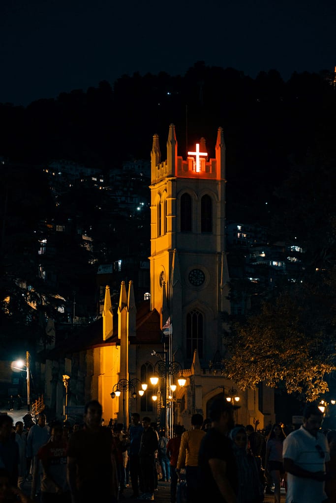 An illuminated church with a glowing red cross surrounded by people in a vibrant urban area at night.